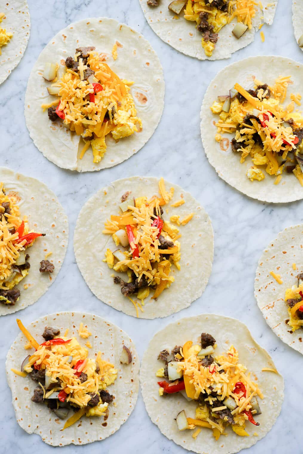 open breakfast tacos filled with scrambled eggs, potatoes, bell peppers, shredded cheese, and crumbed breakfast sausage laying on a marble surface