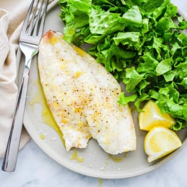 a light gray plate with a filet of red snapper next to a bed of lettuce and lemon wedges
