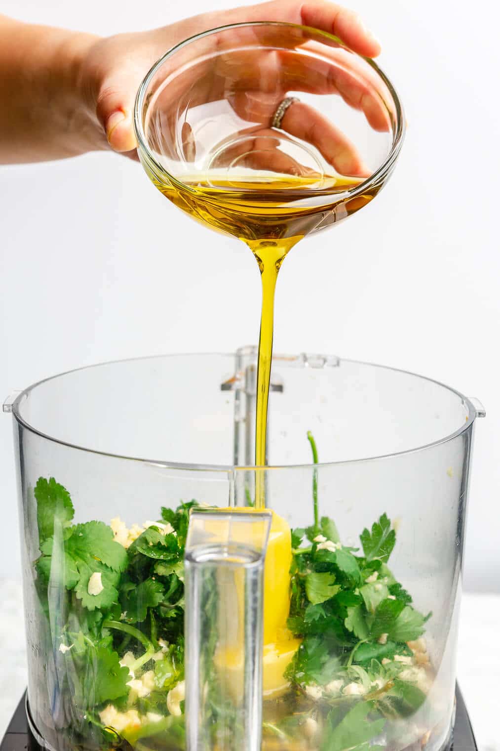Oil being poured into a food processor with herbs, garlic, salt, vinegar, and red pepper flakes.