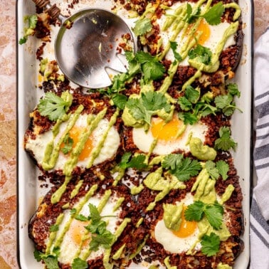 Five, fully prepared, stuffed mushrooms sit on a white baking tray which sits on a pink countertop. A serving spoon sits in place of one of the mushrooms.