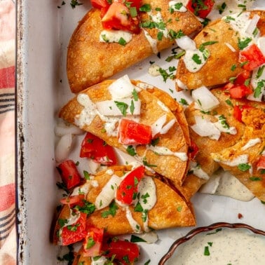 The tacos have been placed. in a white serving tray and have been garnished with diced bell peppers and a white sauce. The remaining sauce sits in a bowl to the side.