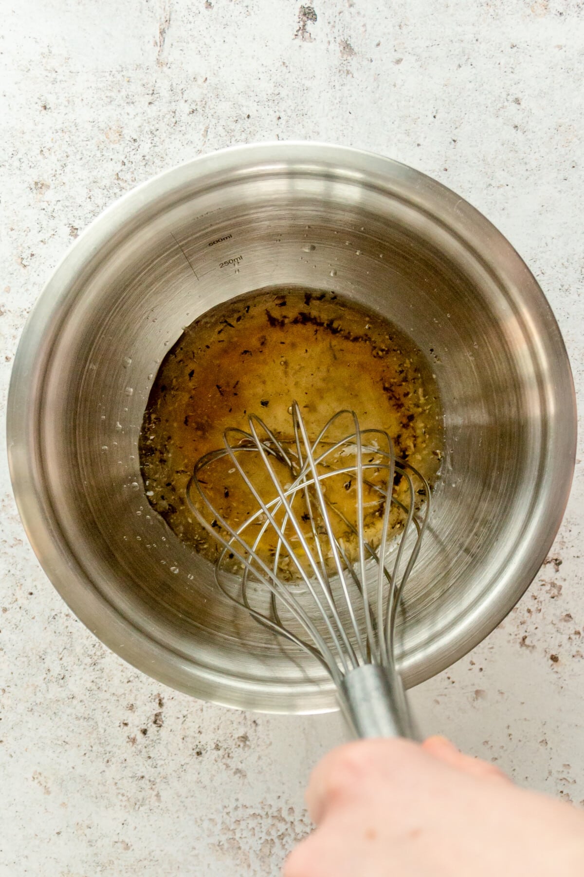 An apple cider vinaigrette is whisked in a stainless steel bowl on a light grey surface.