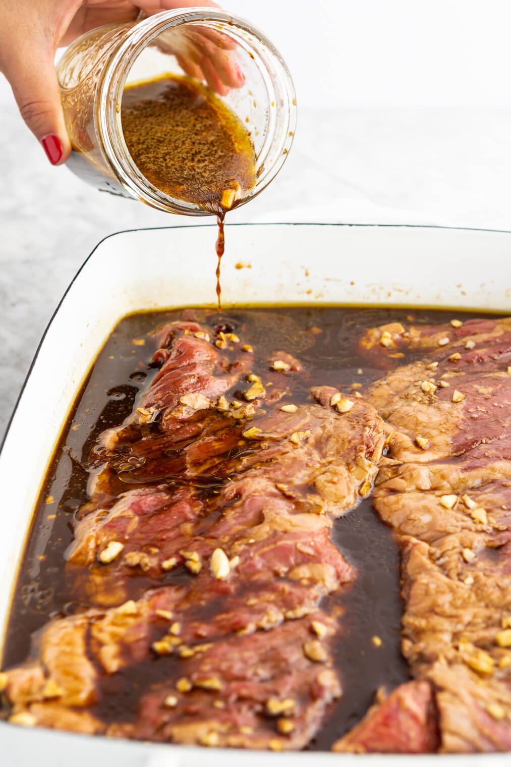 Hand pouring marinade over skirt steak in a casserole dish.